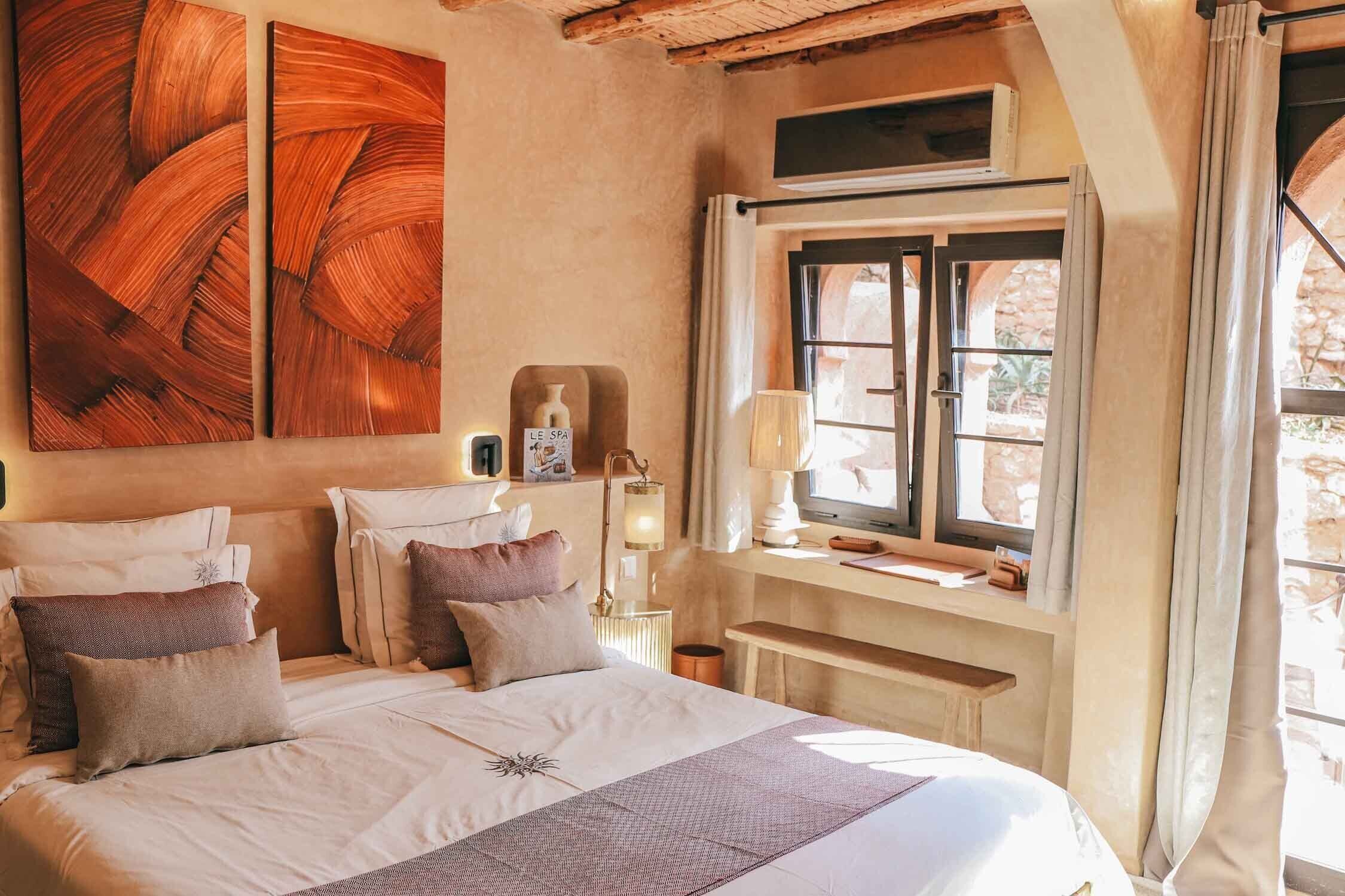 A bedroom with a large bed, a window with curtains, a bedside table with a lamp, and a wall-mounted TV.
