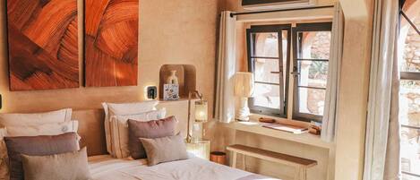 A bedroom with a large bed, a window with curtains, a bedside table with a lamp, and a wall-mounted TV.
