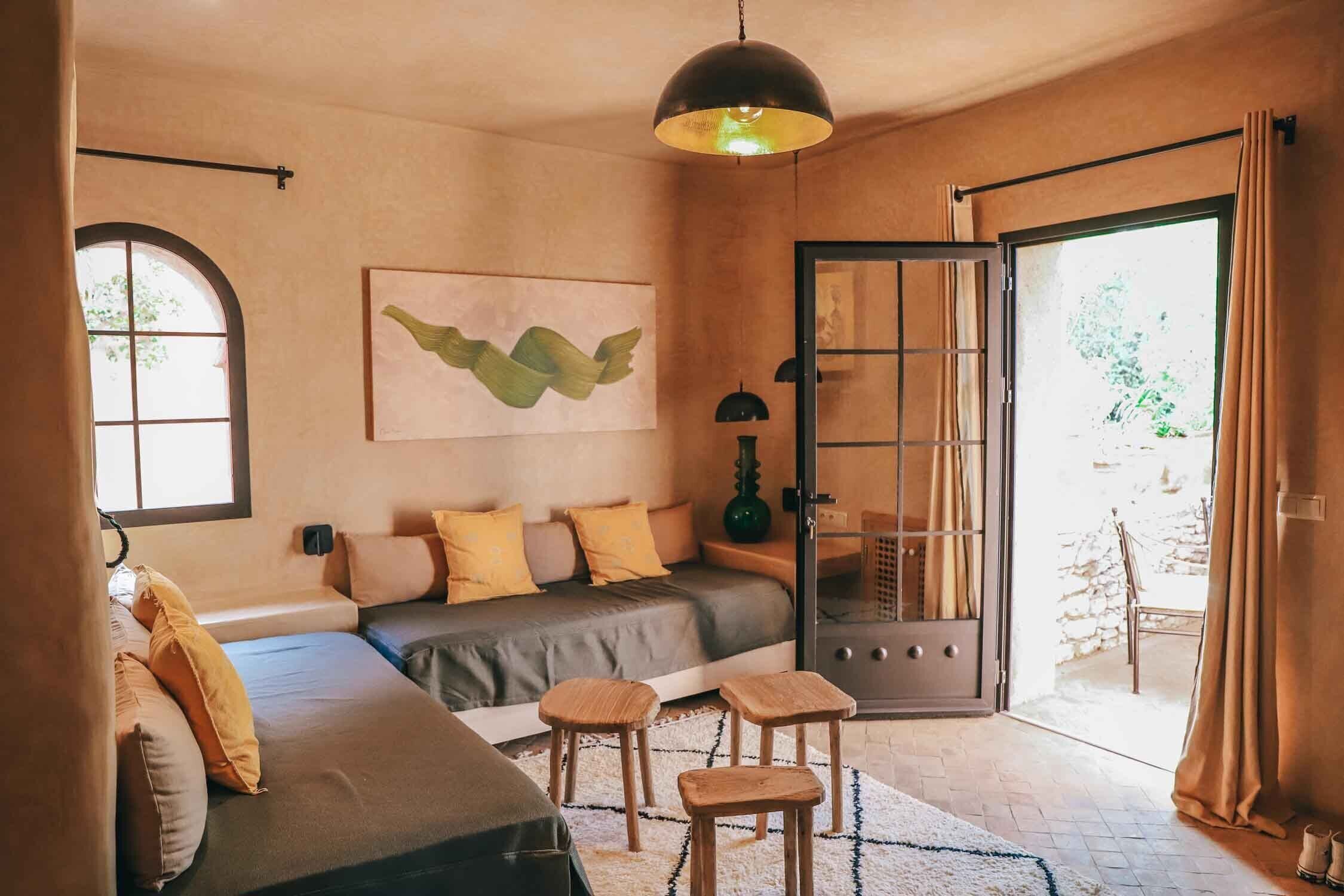 Une pièce chaleureuse avec un canapé, deux tabourets en bois, une grande fenêtre et une porte donnant sur l’extérieur.