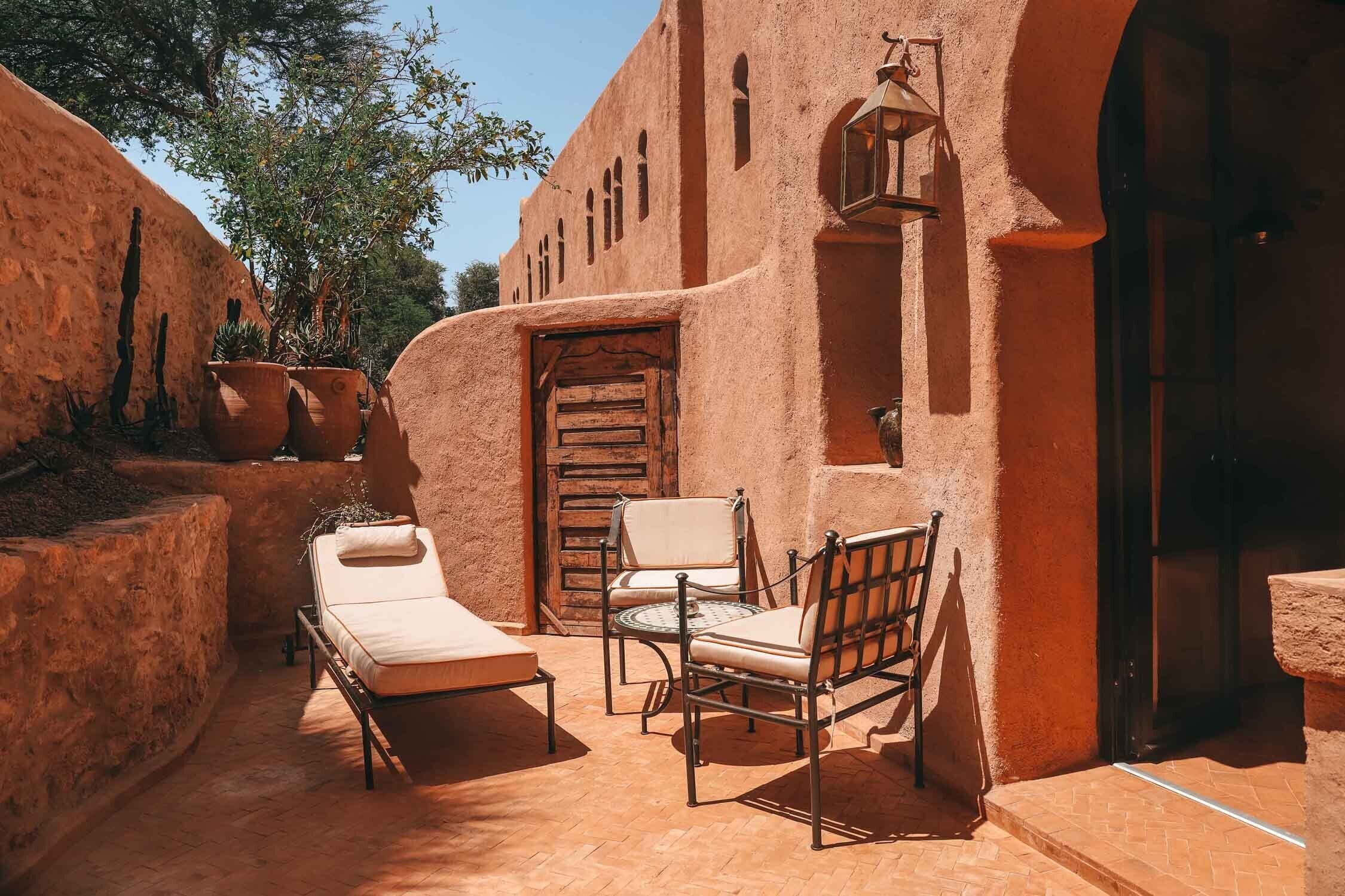 Un espace extérieur ensoleillé, aménagé avec des sièges, comprenant un fauteuil et deux chaises, une porte en bois et une lanterne.