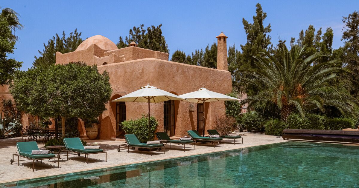 Un espace piscine avec des chaises longues et des parasols, un bâtiment doté d’un dôme et d’une cheminée, et une végétation luxuriante tout autour.