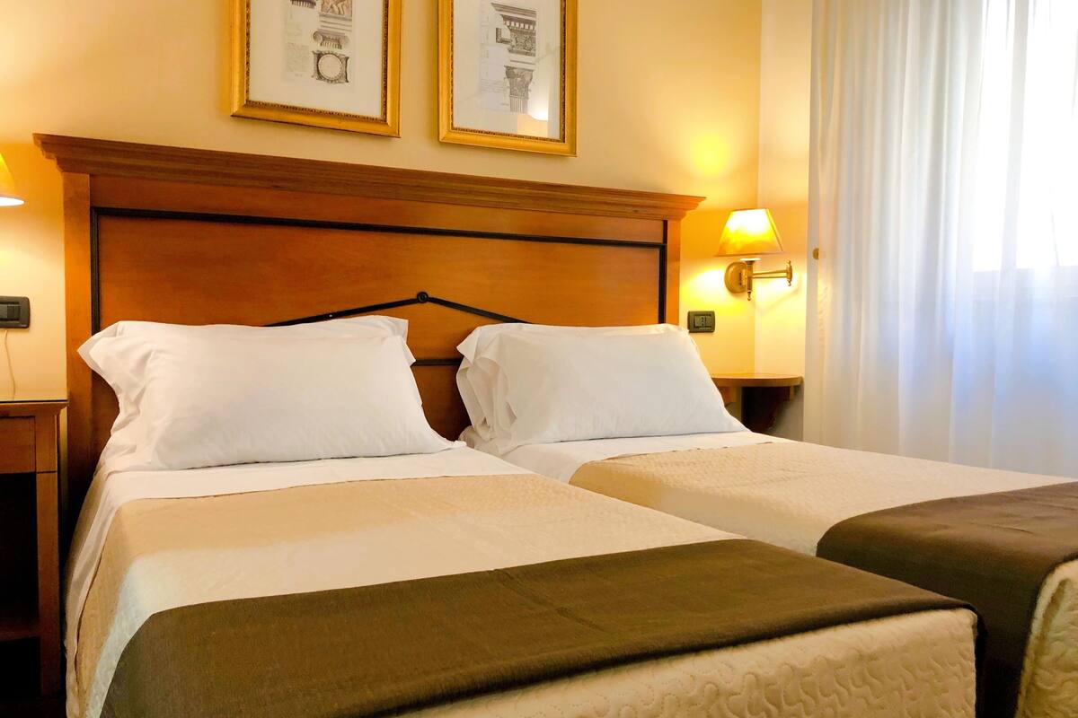 A hotel room with two beds, a wooden headboard, framed pictures on the wall, and a nightstand with a lamp.