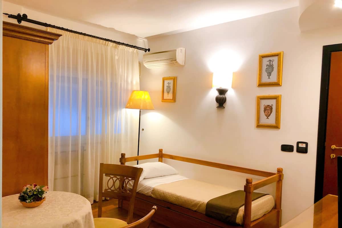 A small, neatly arranged bedroom with a single bed, a chair, a table with a floral centerpiece, and a lamp.