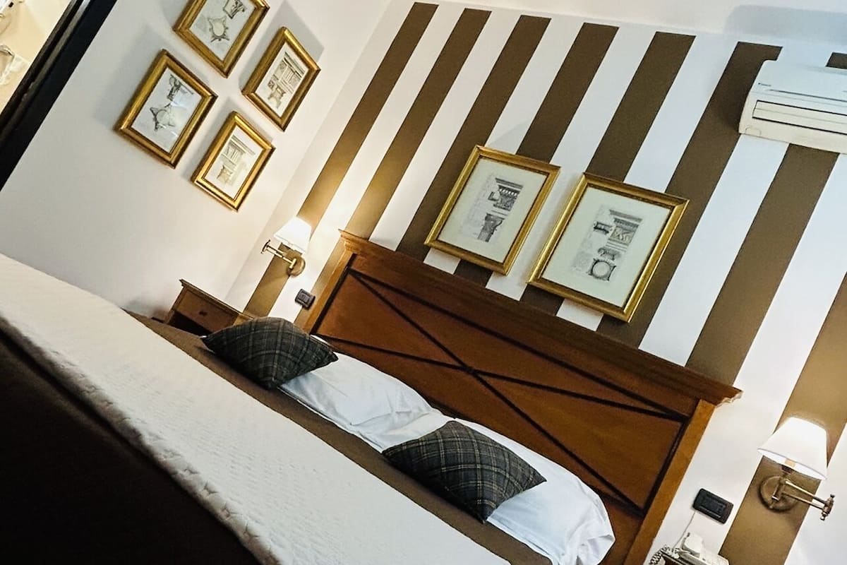 A hotel room with a bed, bedside tables, a wall-mounted air conditioner, and framed pictures on the wall.