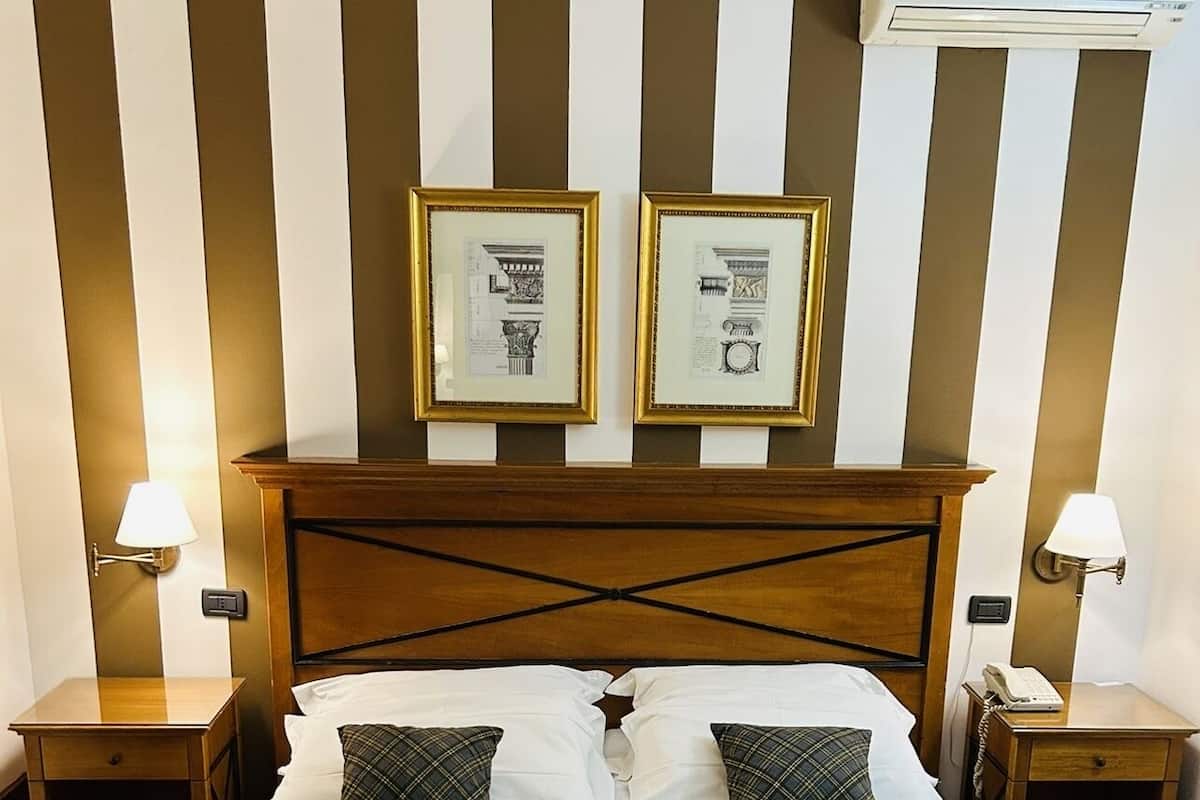 A hotel room with a large bed, two framed pictures on the wall, a wooden headboard, bedside tables with lamps, and an air conditioning unit above the bed.