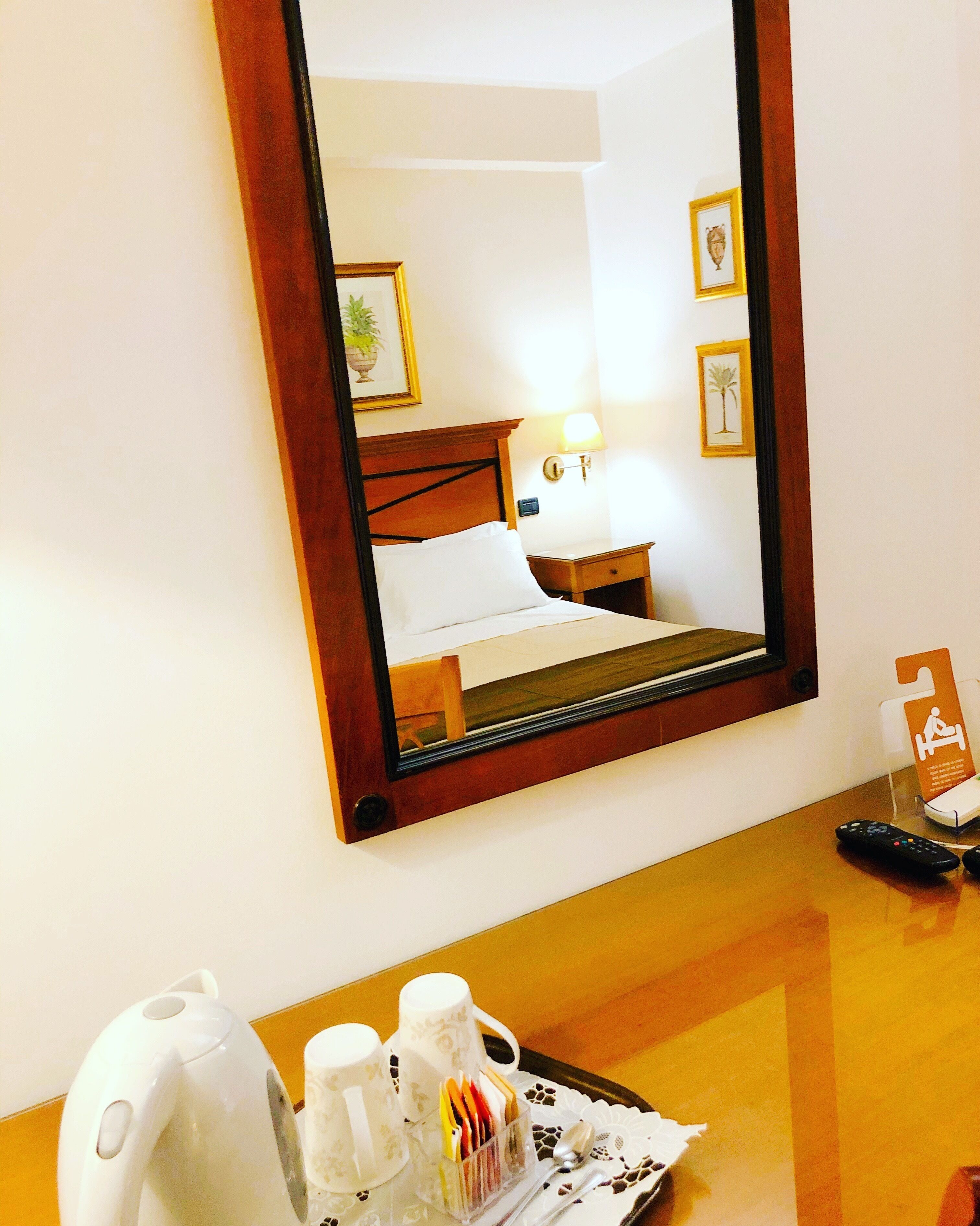 A hotel room with a bed, bedside table, lamp, framed pictures, and a tray with a kettle, cups, and a remote control.
