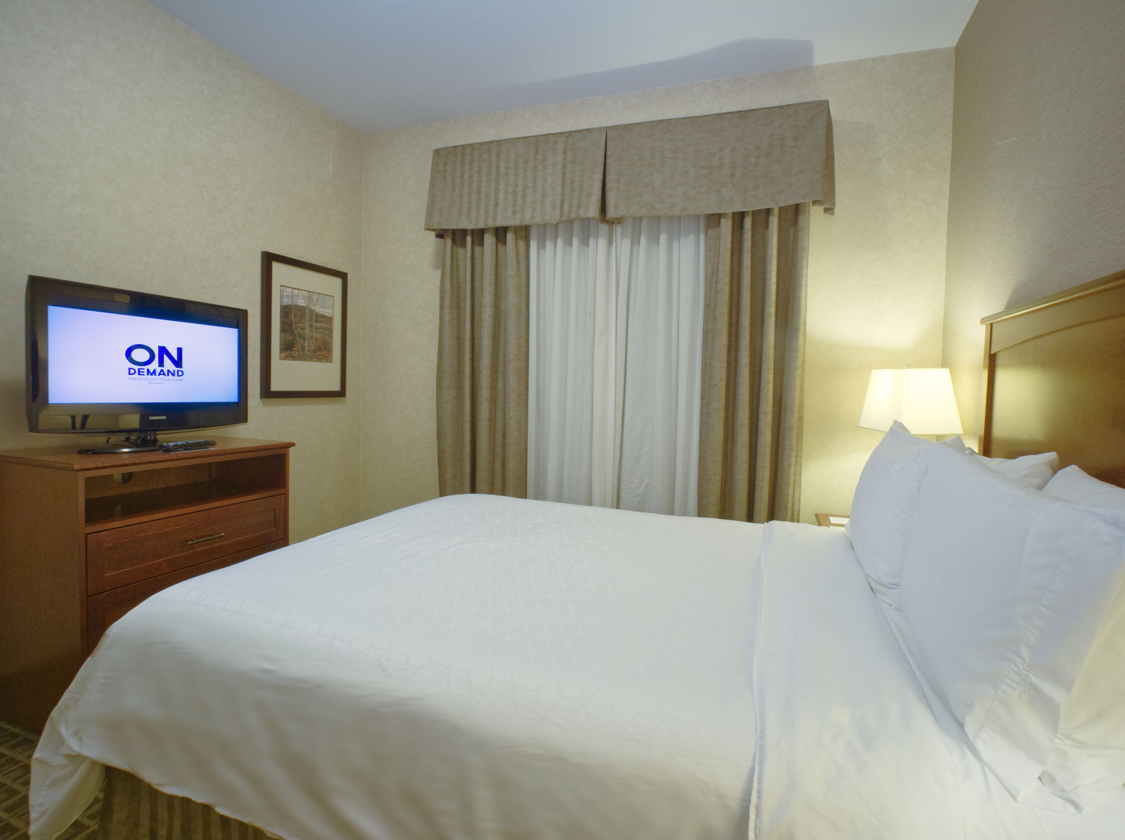 A hotel room with a bed, a television displaying 'ON DEMAND', a lamp, and a window with curtains.