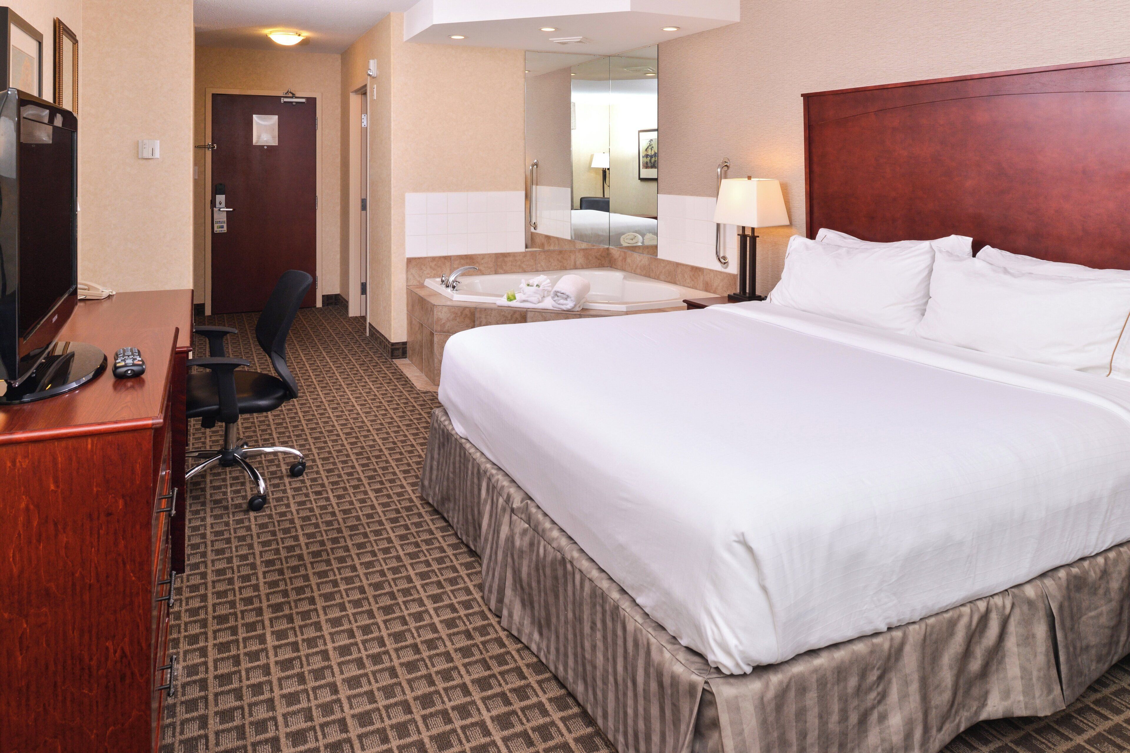 A hotel room with a large bed, a desk with a computer, a chair, a bathroom with a bathtub, and a mirror.