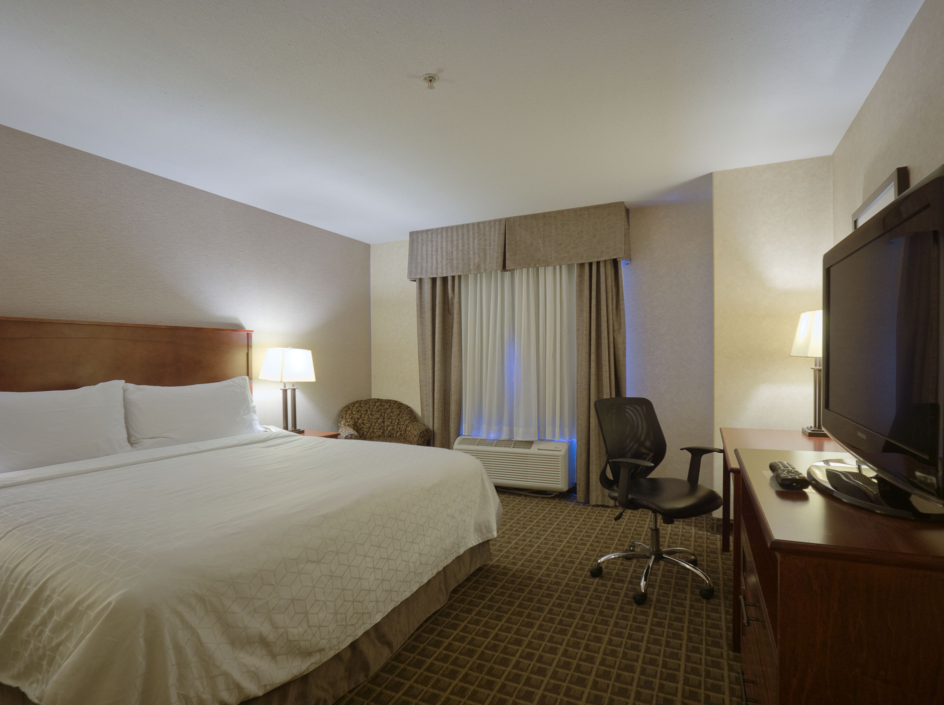 A hotel room with a large bed, a desk with a television, a chair, a lamp, and a window with curtains.