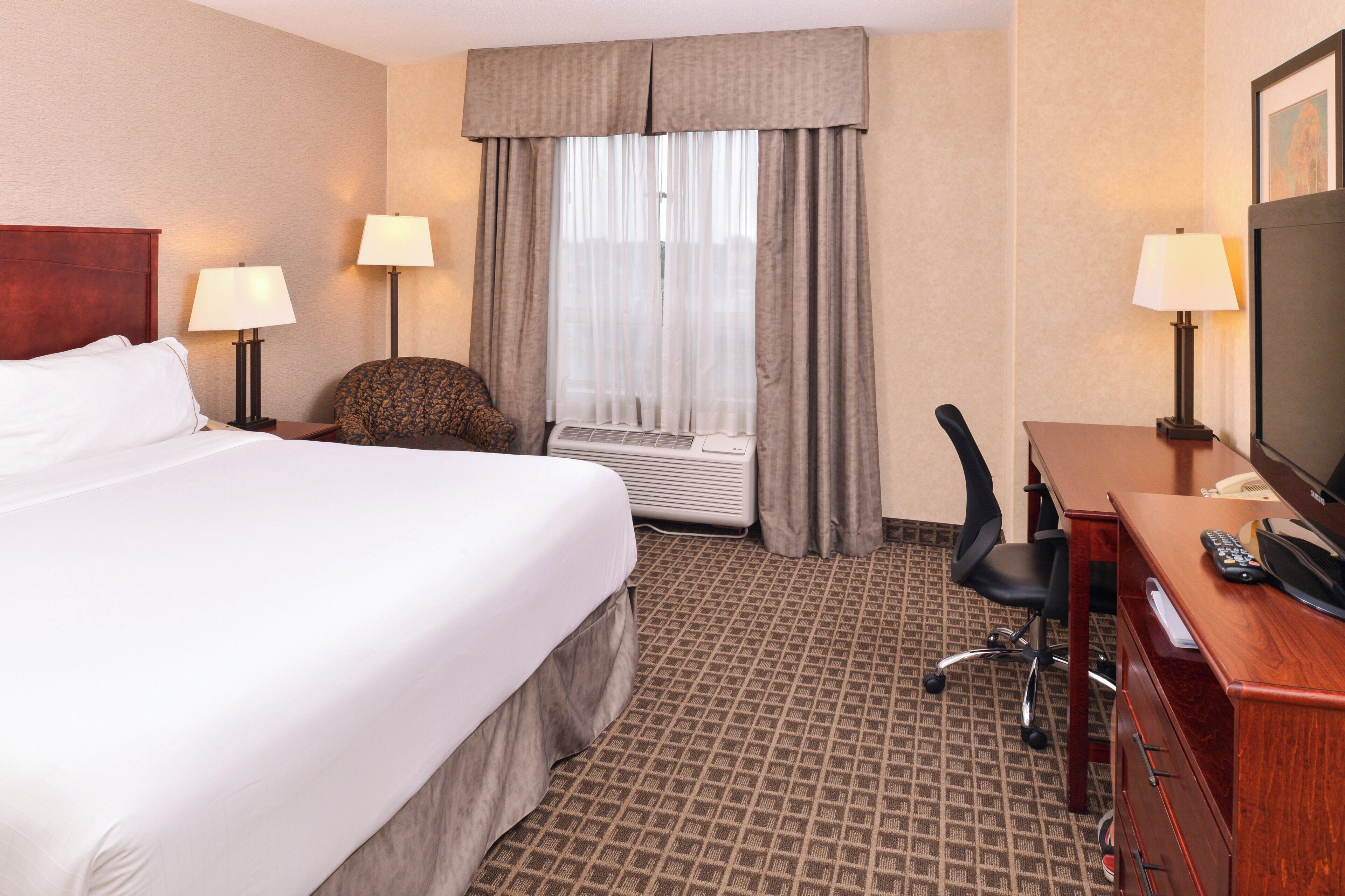 Habitación de hotel con una cama grande, un escritorio con televisor, una silla y una ventana con cortinas.