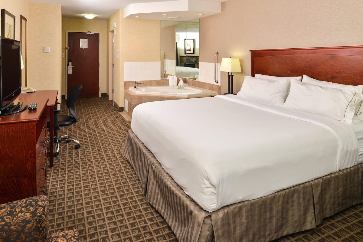A hotel room with a large bed, a desk with a computer, a chair, a bathroom with a sink and mirror, and a television.