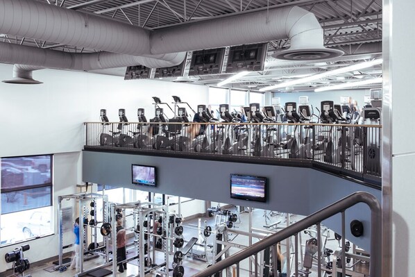 Une salle de sport à plusieurs niveaux, équipée de tapis de course, d’appareils de musculation et d’une mezzanine où se trouve du matériel d’exercice.