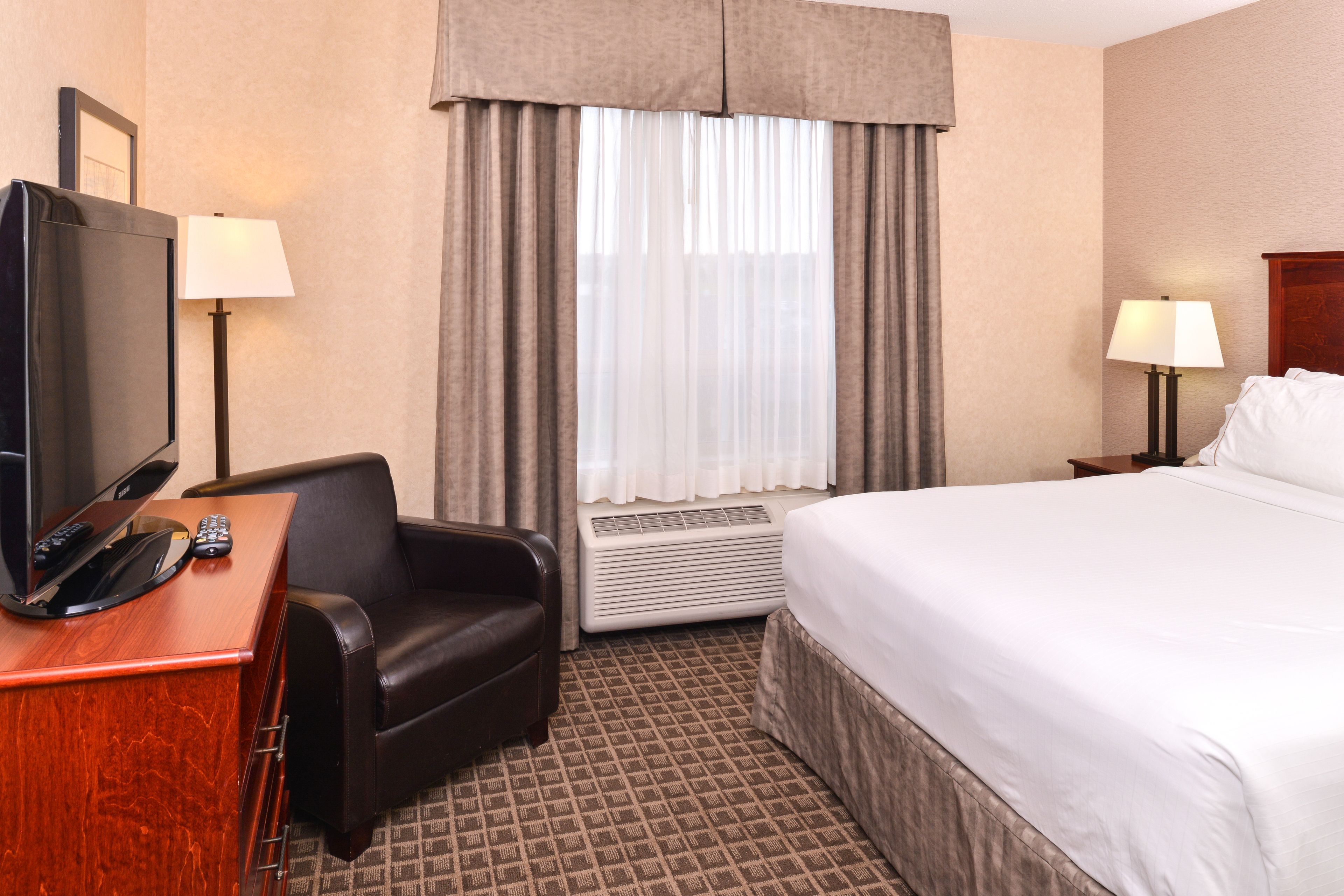 Une chambre d’hôtel comprenant un lit, un bureau avec une télévision et une télécommande, une chaise, une lampe et une fenêtre avec des rideaux.