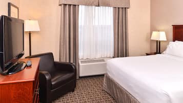 Une chambre d’hôtel comprenant un lit, un bureau avec une télévision et une télécommande, une chaise, une lampe et une fenêtre avec des rideaux.