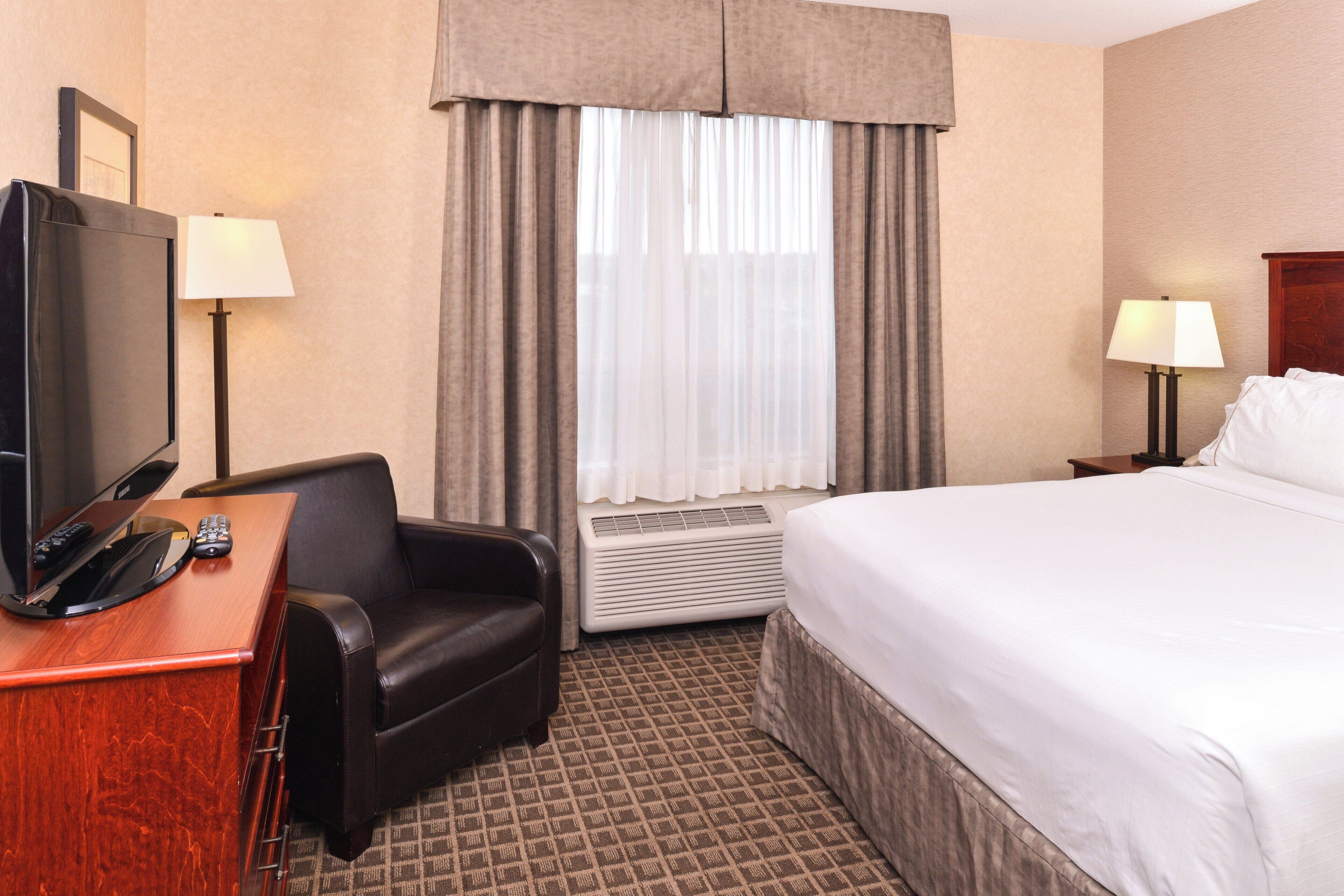 Une chambre d’hôtel comprenant un lit, un bureau avec une télévision, une chaise, une table de chevet avec une lampe et une fenêtre avec des rideaux.