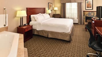 A hotel room with a large bed, a desk with a computer, a chair, a desk lamp, a bedside table, a TV, and a bathroom with a bathtub.