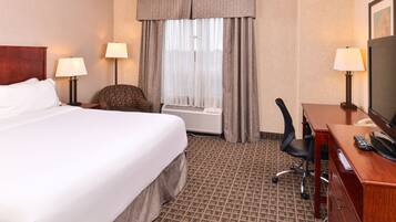 A hotel room with a large bed, two bedside lamps, a desk with a chair, a television, and an air conditioning unit.