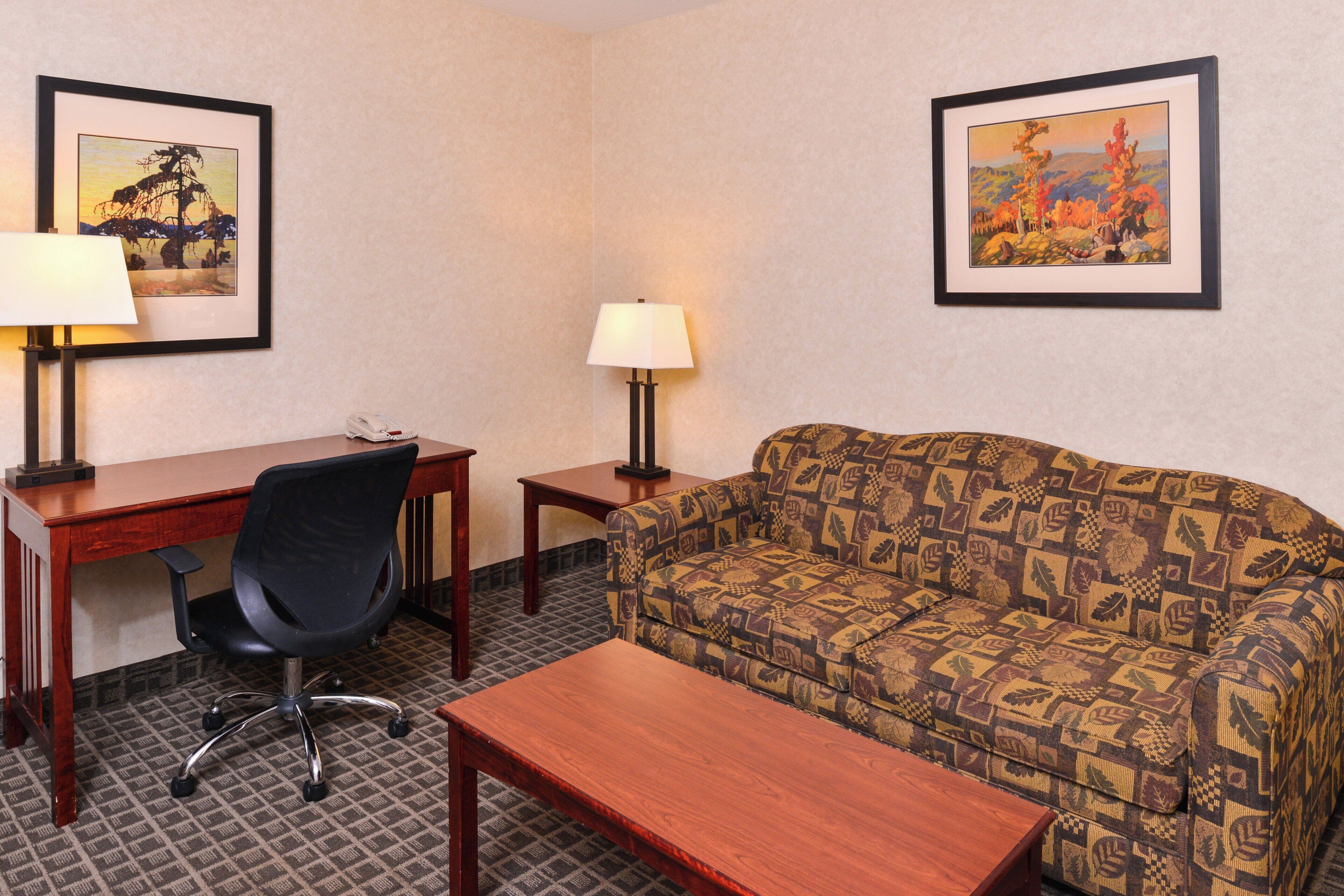 A hotel room with a wooden desk, a black office chair, a patterned sofa, a small table, two lamps, and two framed pictures on the walls.