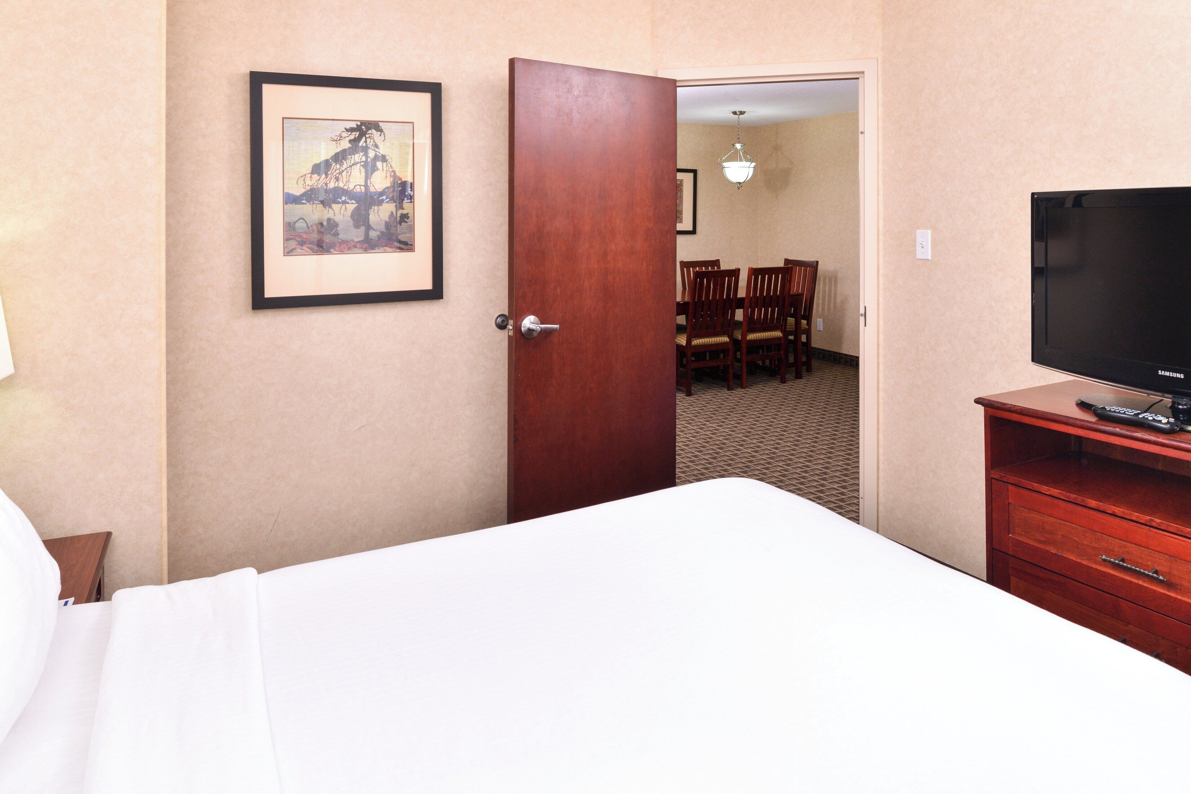 A hotel room with a bed, a TV, a wooden dresser, a framed picture on the wall, and a dining area visible through an open door.