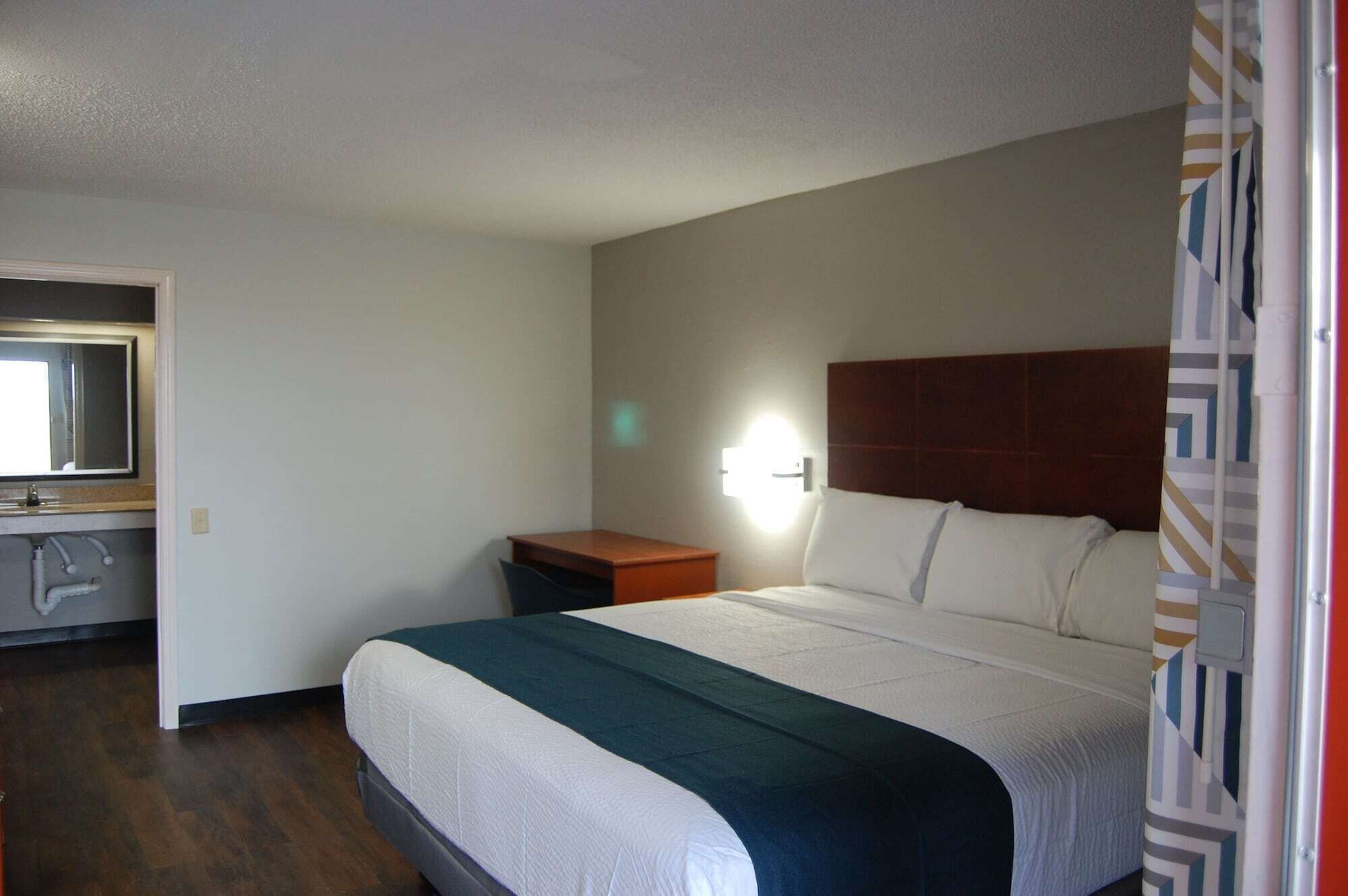 A hotel room with a bed, a nightstand, a mirror, and a wall-mounted light.