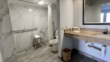 A bathroom with a marble shower, a toilet, a sink with a mirror, and a trash can.