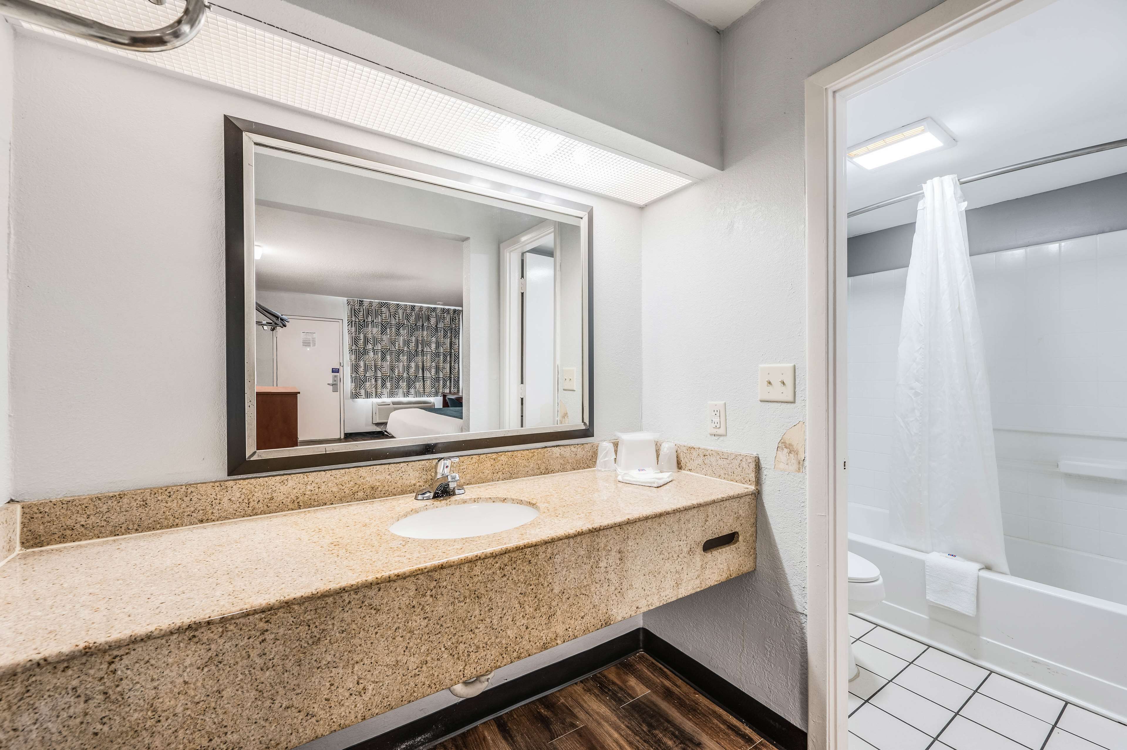 Une salle de bain avec un lavabo surmonté d’un plan de travail en granit, un miroir, une douche avec un rideau blanc et un sol carrelé.
