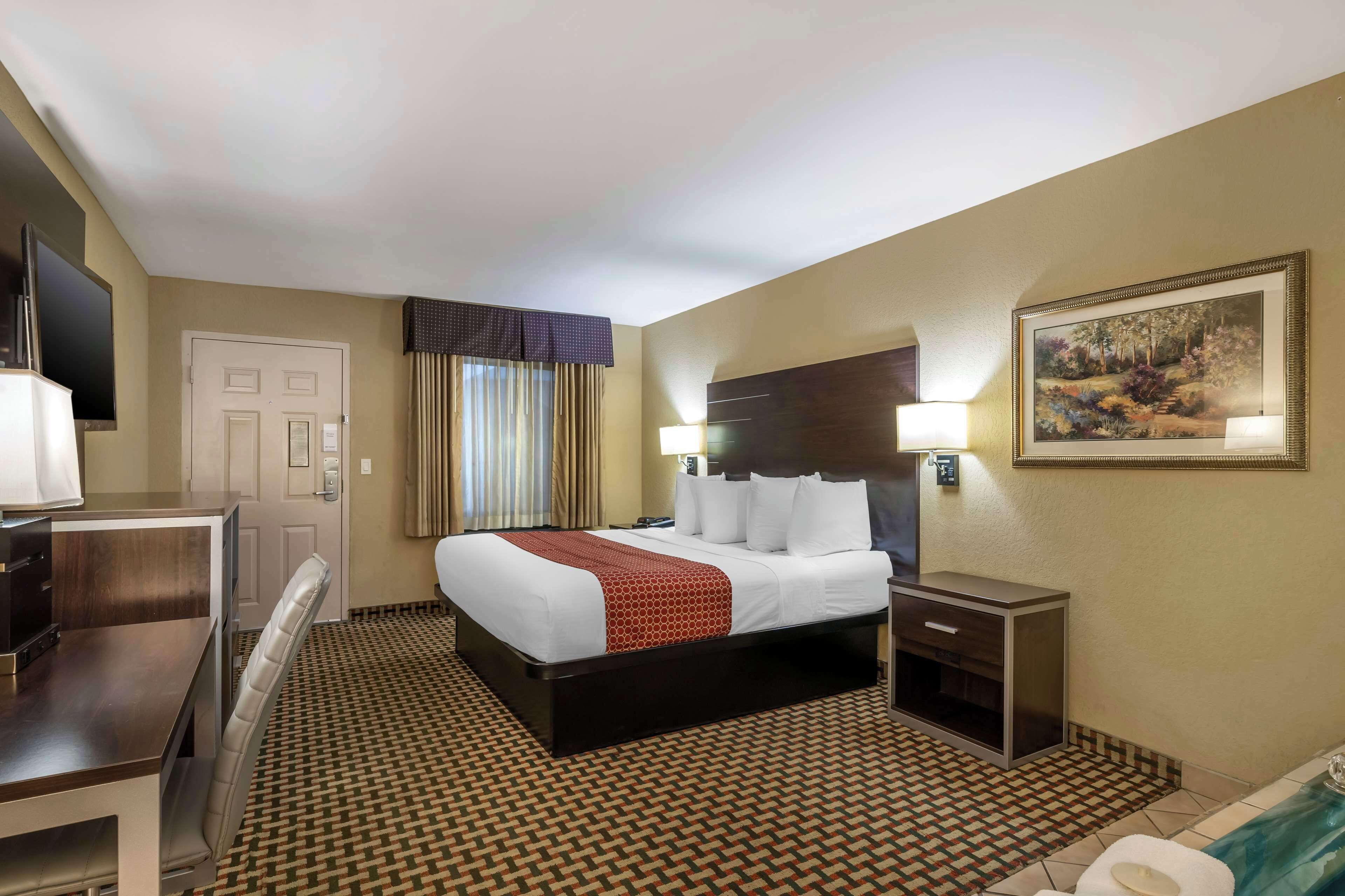 Habitación de hotel con cama, escritorio, silla, mesita de noche, lámpara, cuadro y baño con inodoro y lavamanos.