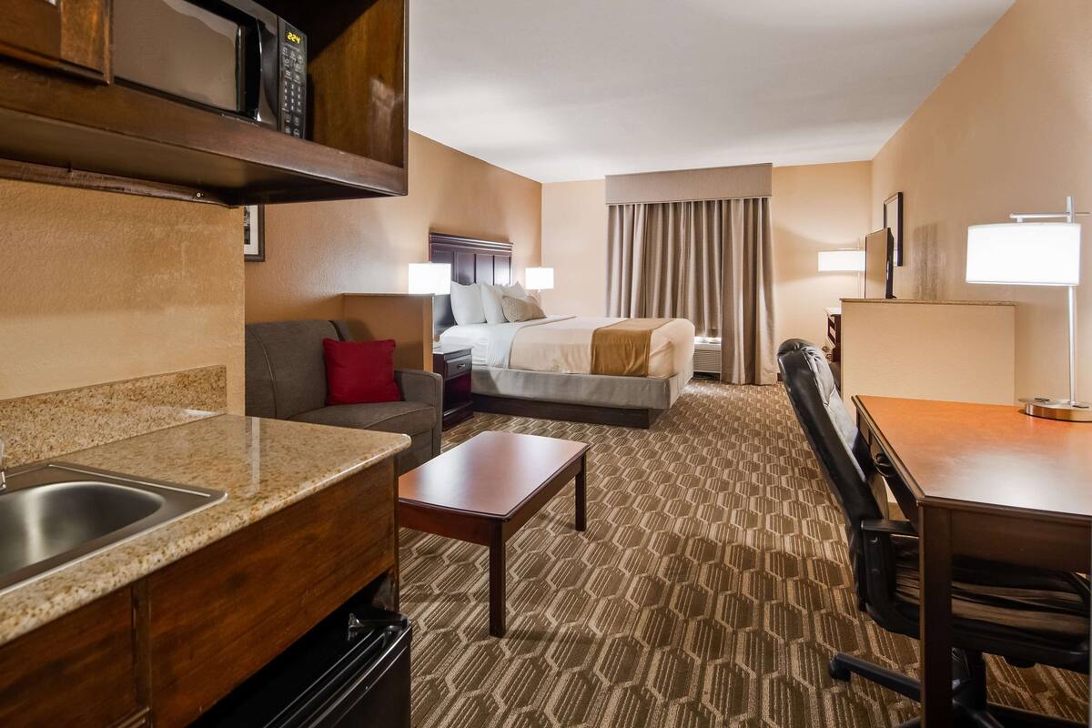 A hotel room with a kitchenette, a bed, a desk, and a seating area.