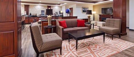 A modern living room with a sofa, two armchairs, a coffee table, and a patterned rug. A kitchen area with a stone wall and various appliances is visible in the background.