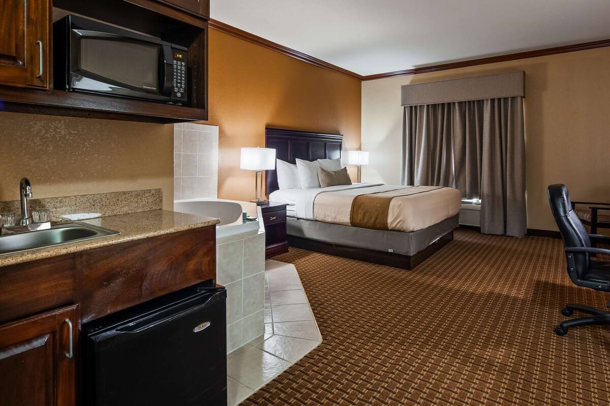 A hotel room with a kitchenette, a bed with a headboard, a desk with a chair, and a bathroom with a shower and toilet.