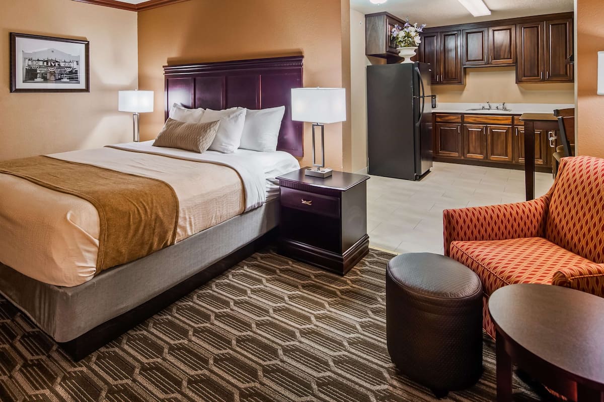 A hotel room with a bed, a nightstand, a chair, a small table, and a kitchen area.