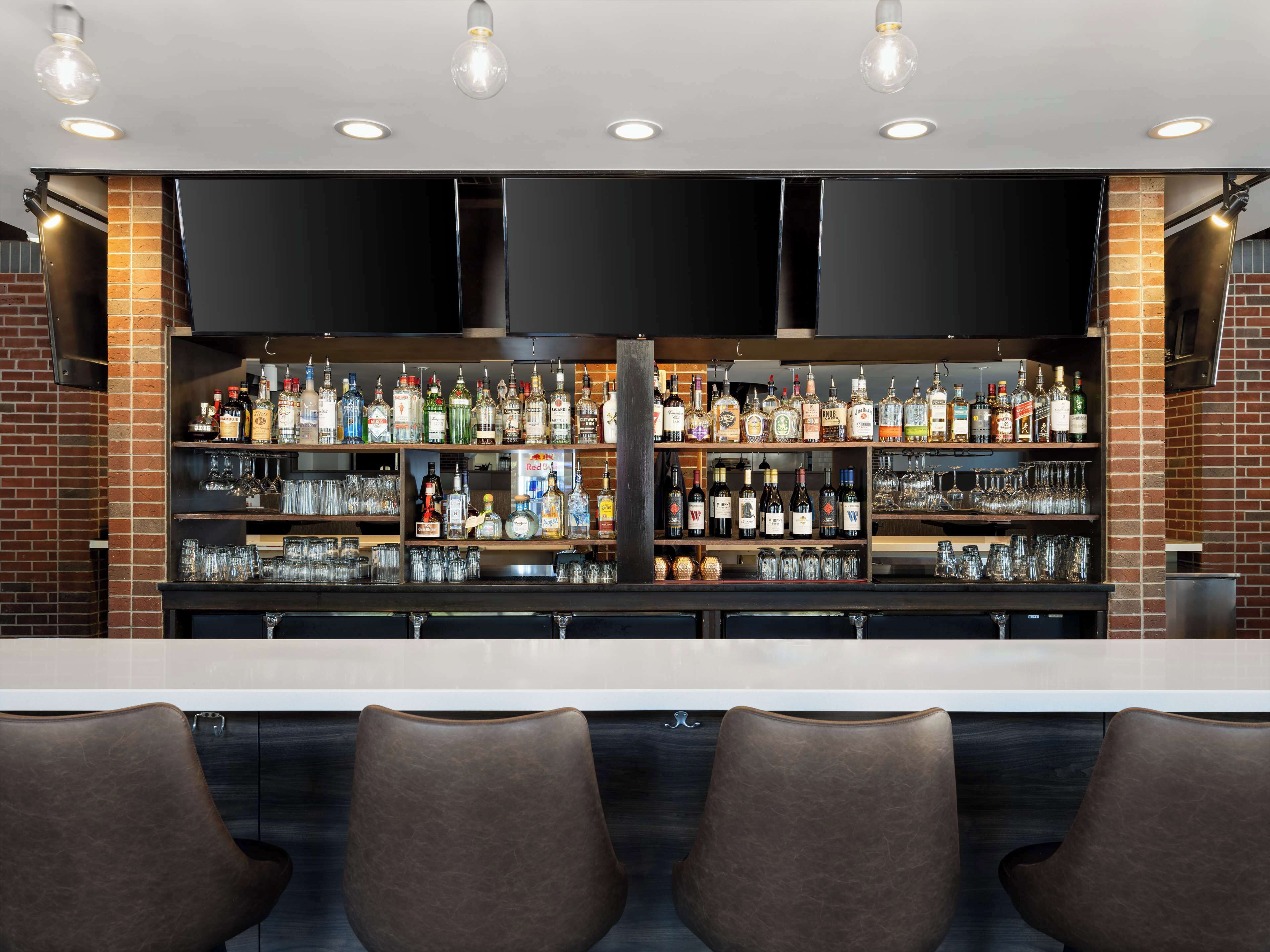 Un bar con varias estanterías llenas de botellas de licor y vasos, un televisor de pantalla plana y taburetes de cuero.