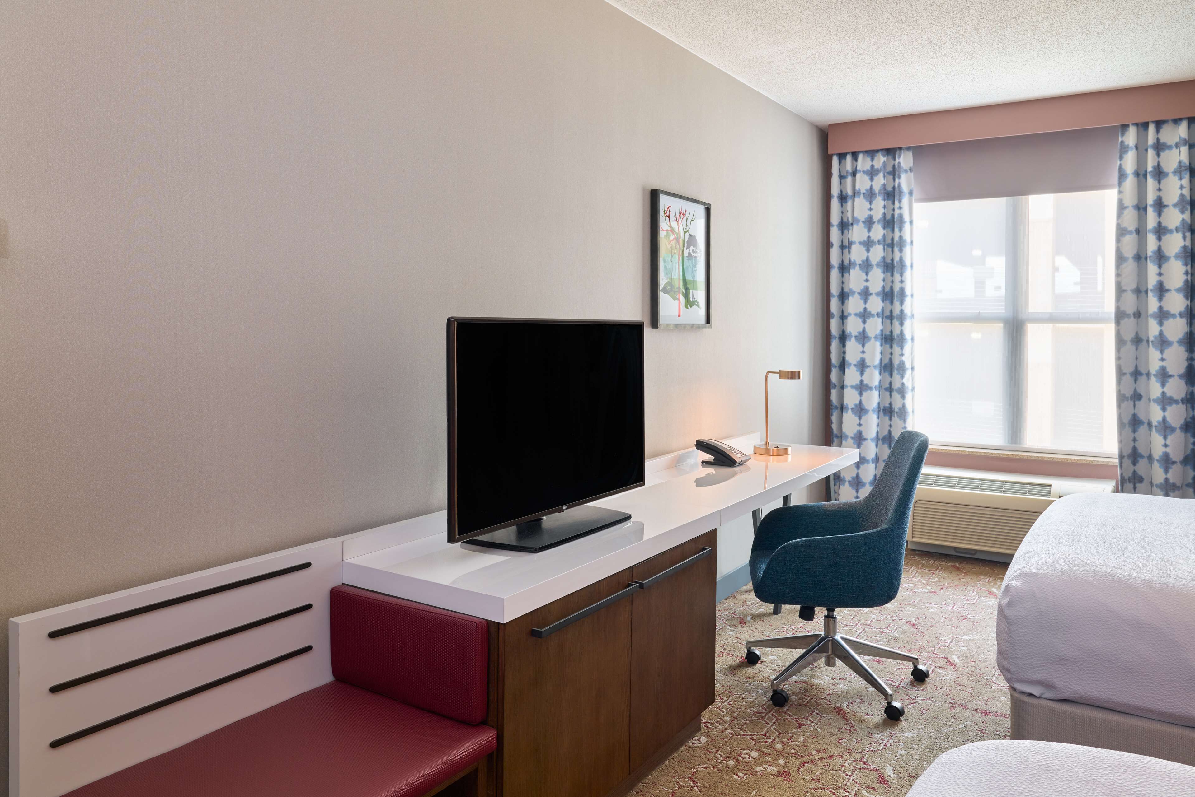 Une chambre d’hôtel avec un lit, un bureau avec une télévision, une chaise et une fenêtre avec des rideaux.
