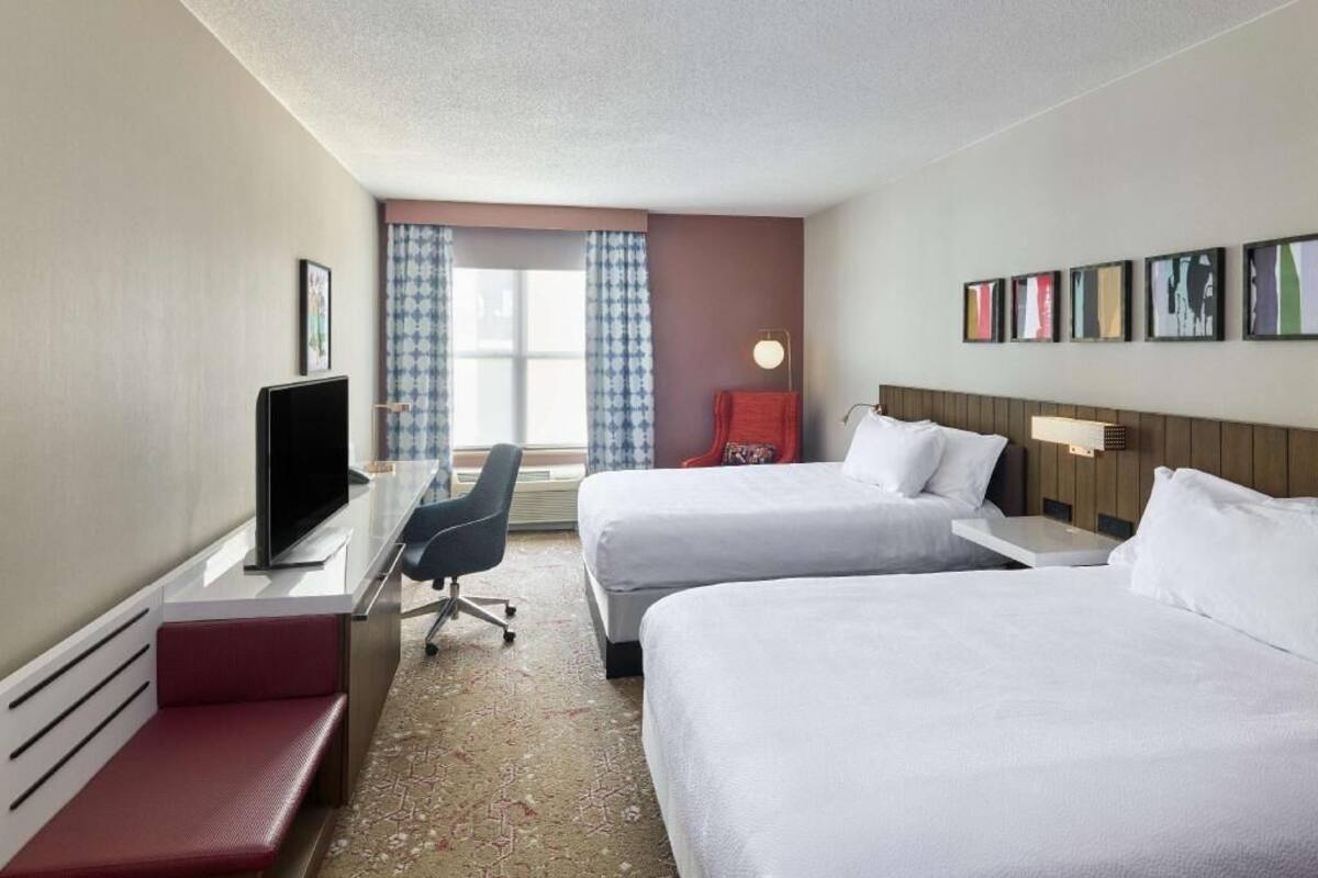 A hotel room with two beds, a desk with a TV, a chair, a window with curtains, and framed pictures on the wall.