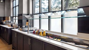A modern kitchen with a glass-top buffet, stainless steel appliances, and a view of trees through large windows. - Hilton Garden Inn Bloomington (Bloomington)