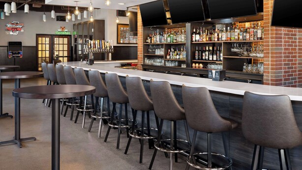 A bar area with a long counter, multiple stools, and a variety of liquor bottles and glasses. - Hilton Garden Inn Bloomington (Bloomington)