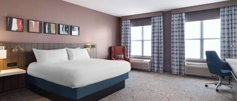 A hotel room with a large bed, a desk, a chair, a window with curtains, and wall-mounted artwork.