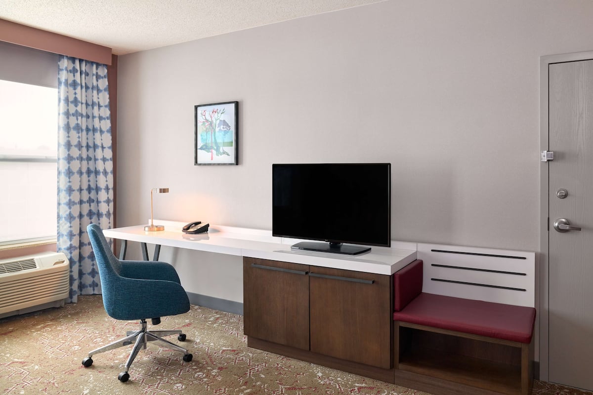 A hotel room with a desk, a television, a chair, a bench, a picture on the wall, and a door.