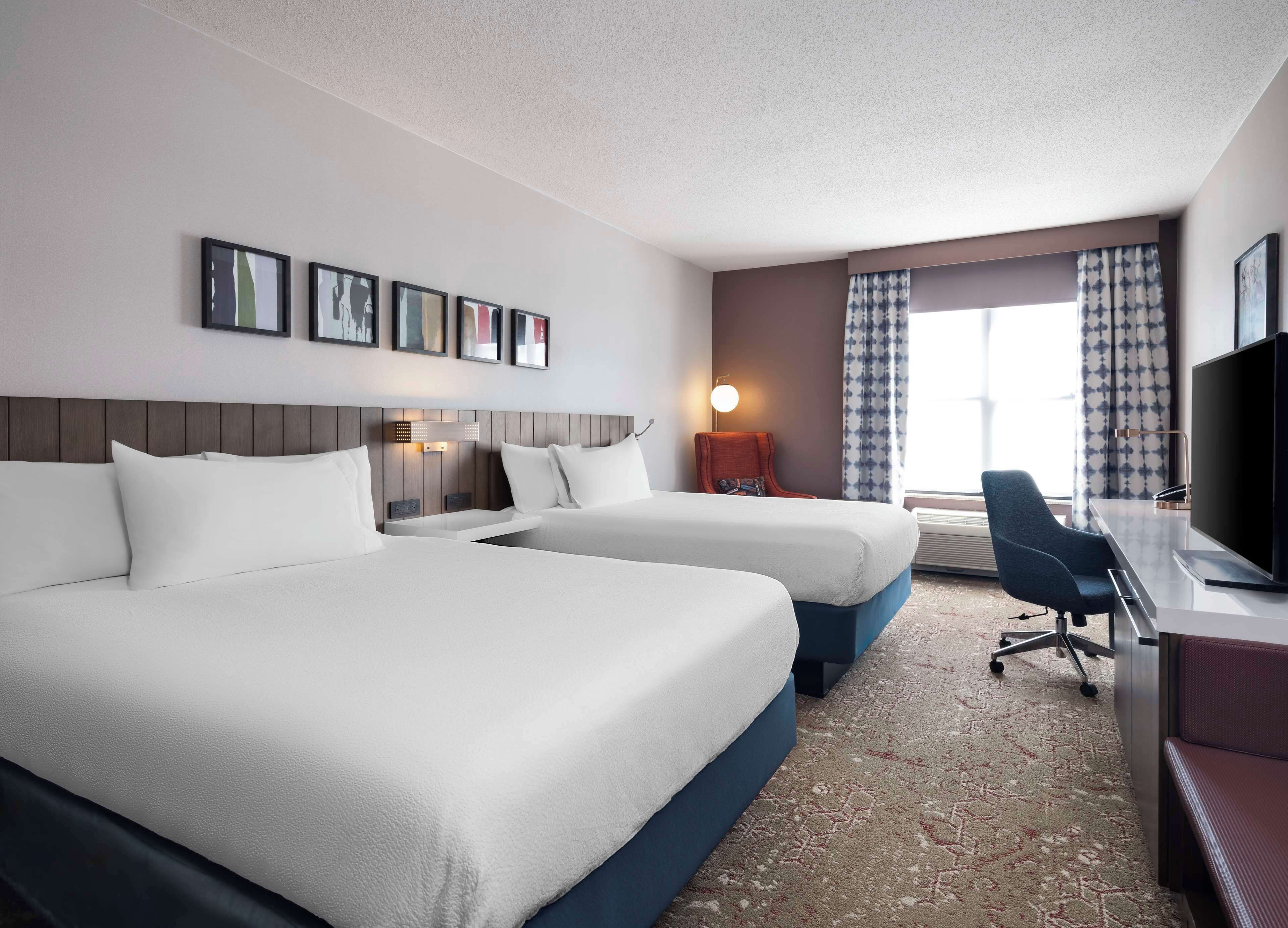 A hotel room with two beds, a desk with a TV, a chair, and a window with curtains.