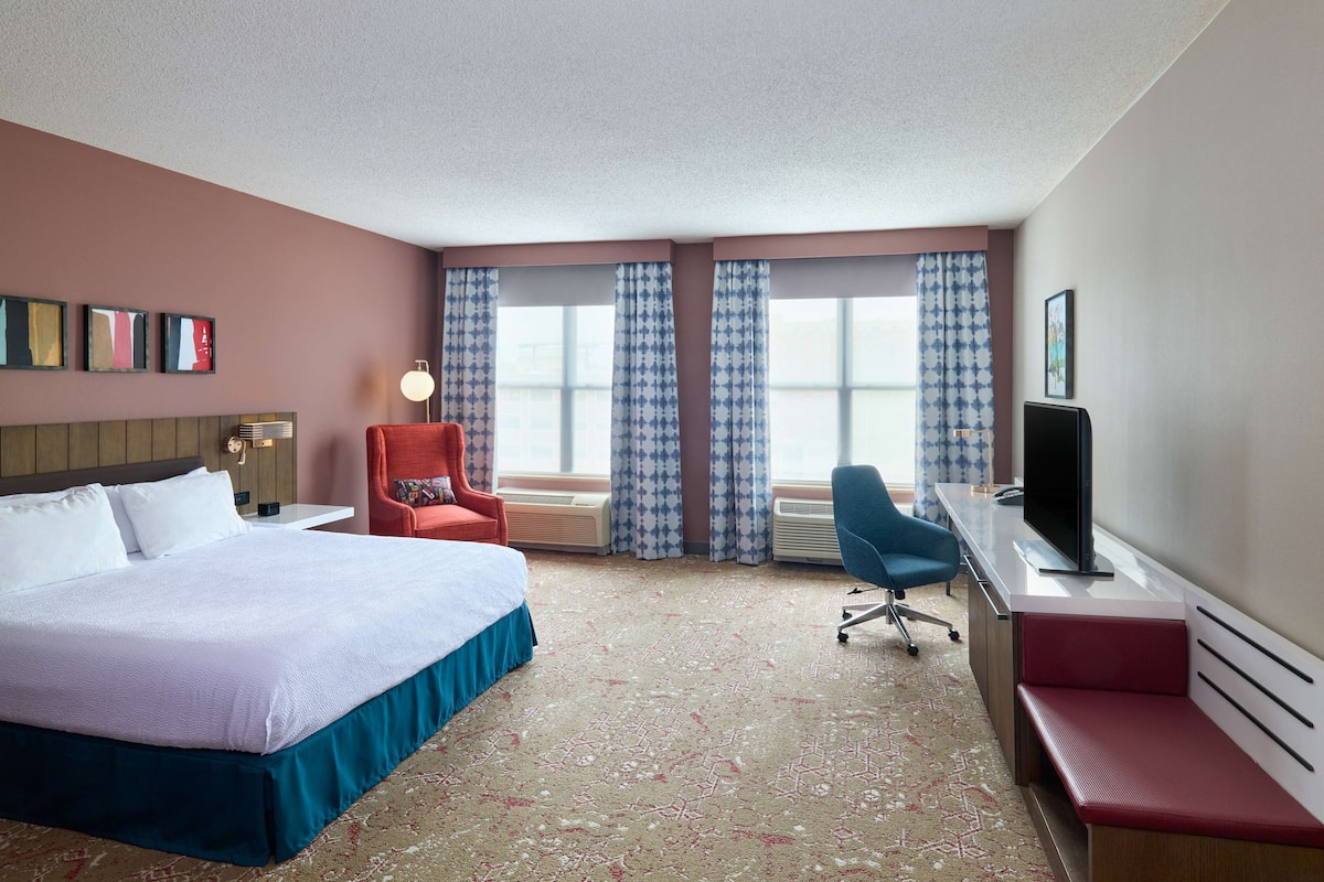 A hotel room with a bed, a desk, a chair, a TV, and a window with patterned curtains.