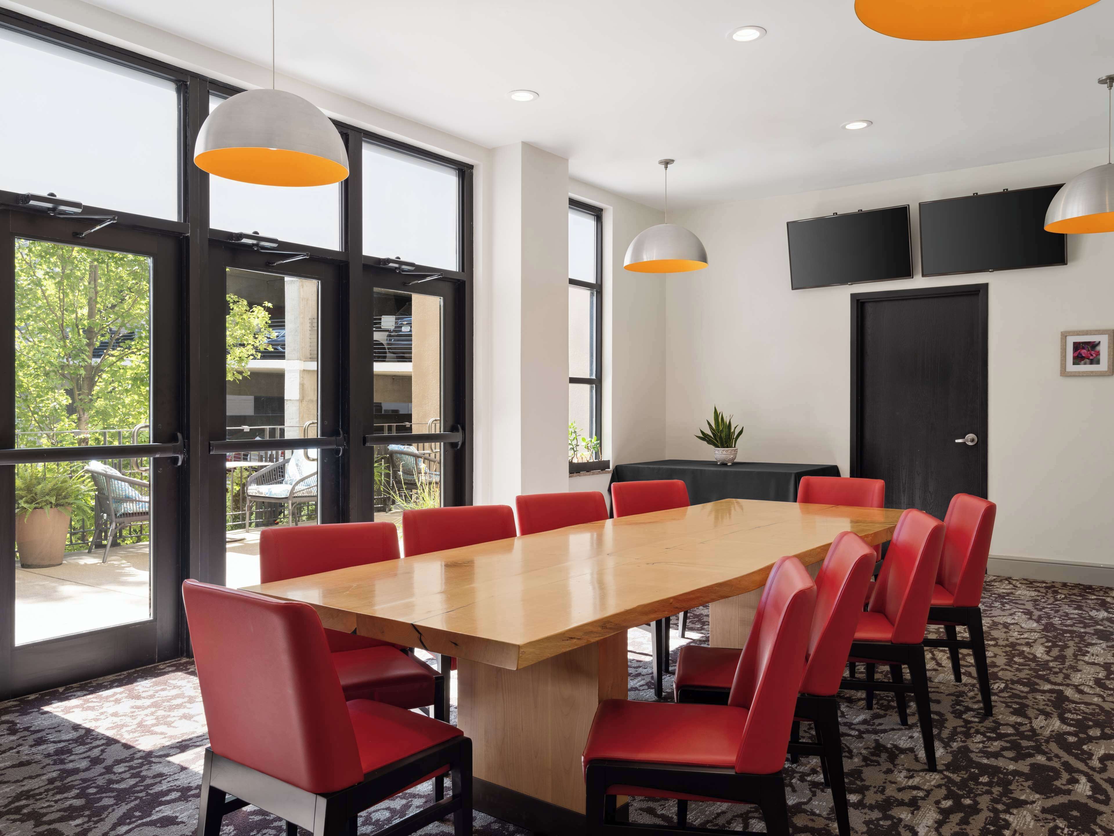 A conference room with a long table, red chairs, large windows, and ceiling-mounted lights.