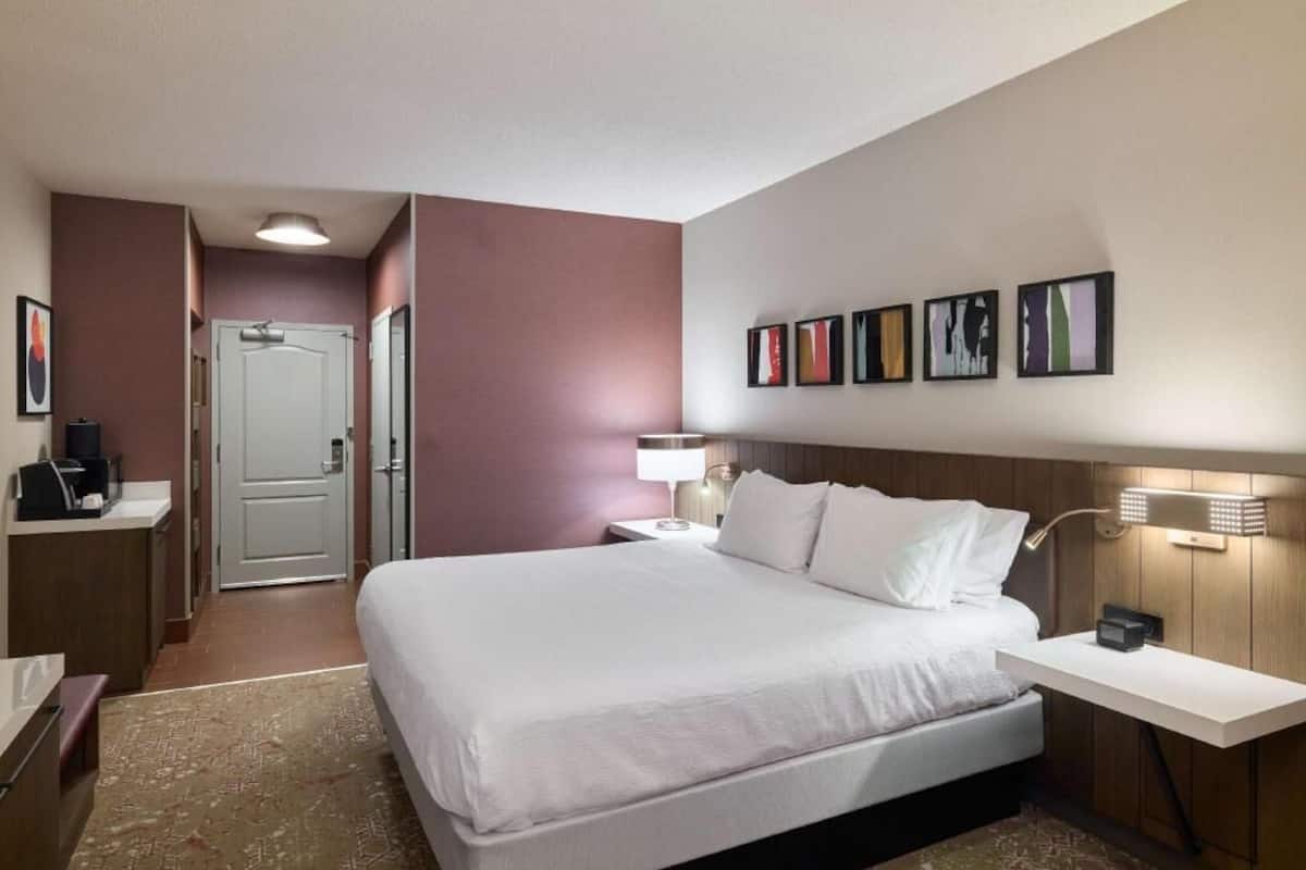 A hotel room with a bed, bedside table, lamp, artwork on the wall, and a door leading to another room.