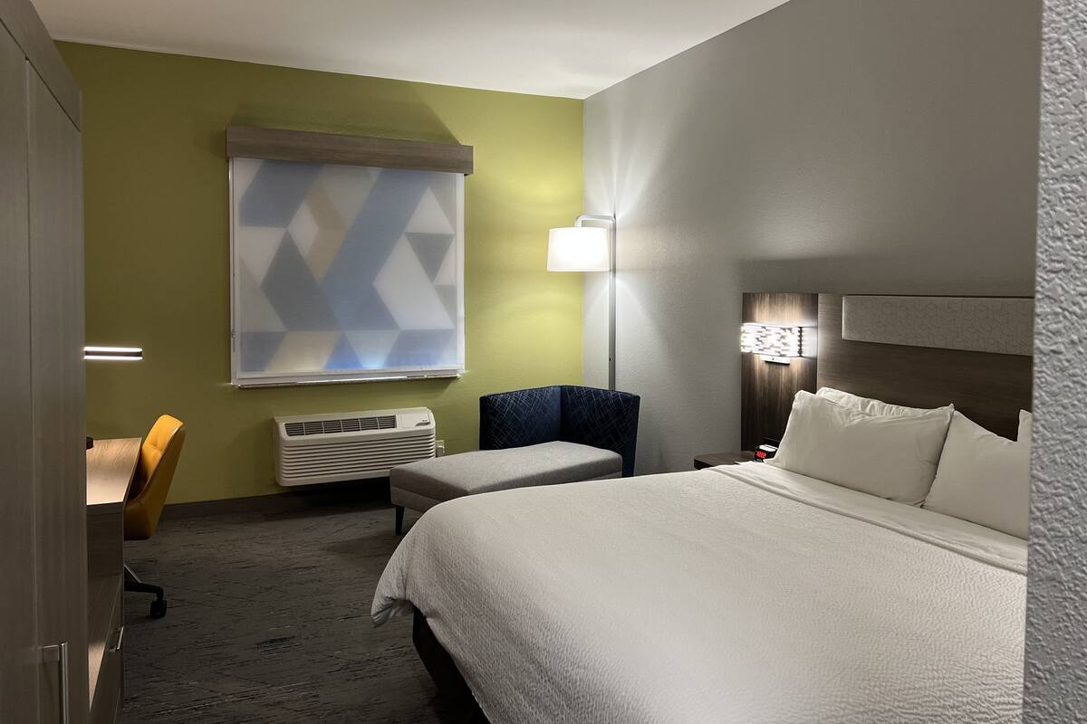 A hotel room with a large bed, a desk, a chair, and a window with a geometric pattern.