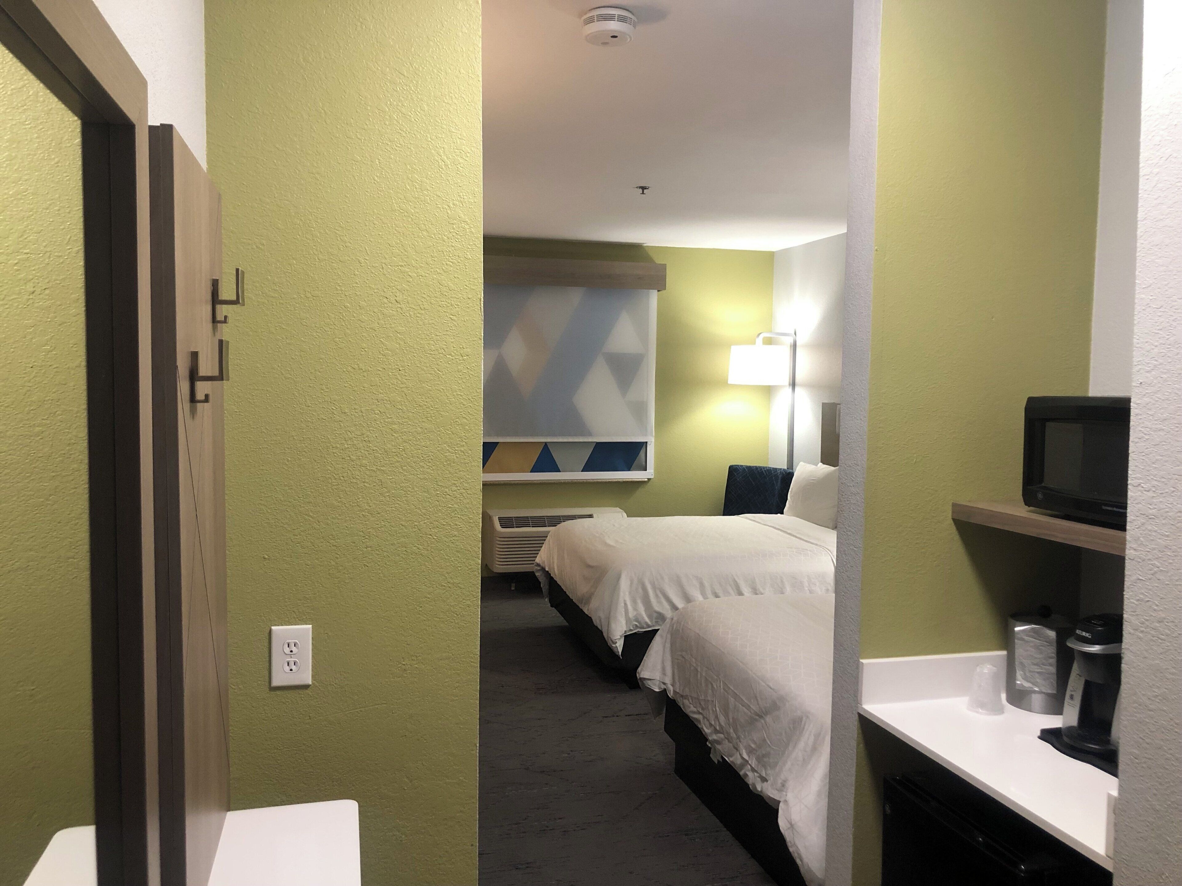 Una habitación de hotel con cama, mesita de noche, lámpara y un microondas en un estante.