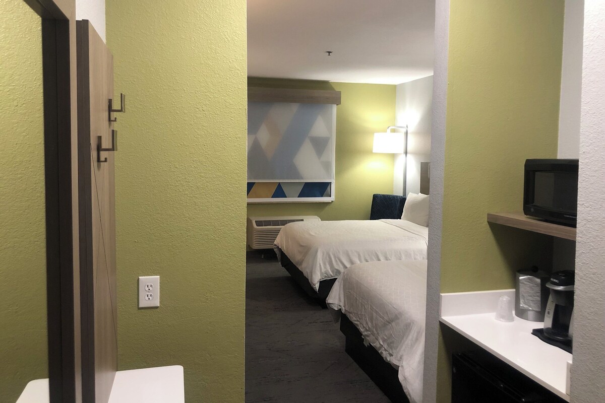 A hotel room with a bed, bedside table, lamp, and a microwave on a shelf.