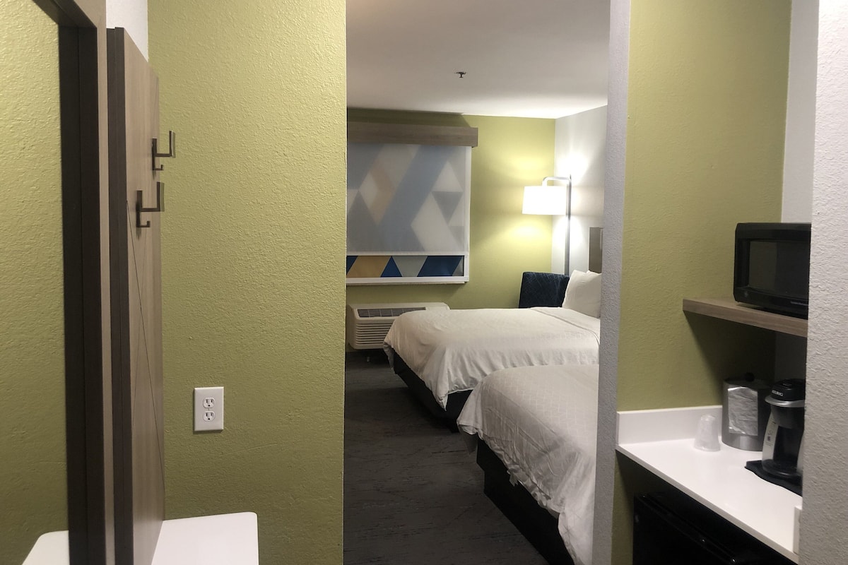 A hotel room with a bed, bedside table, lamp, and a microwave on a shelf.