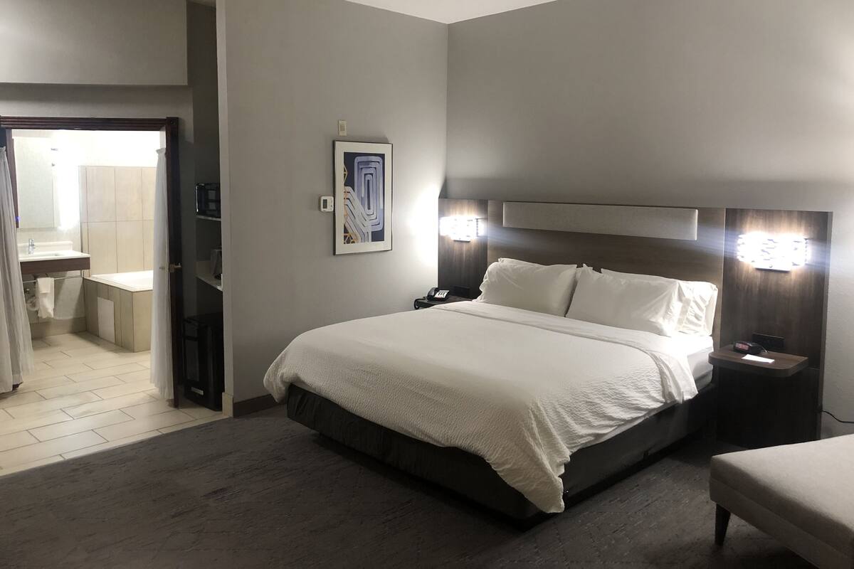 A hotel room with a bed, bedside table, and a nightstand. There is a bathroom visible through an open door.