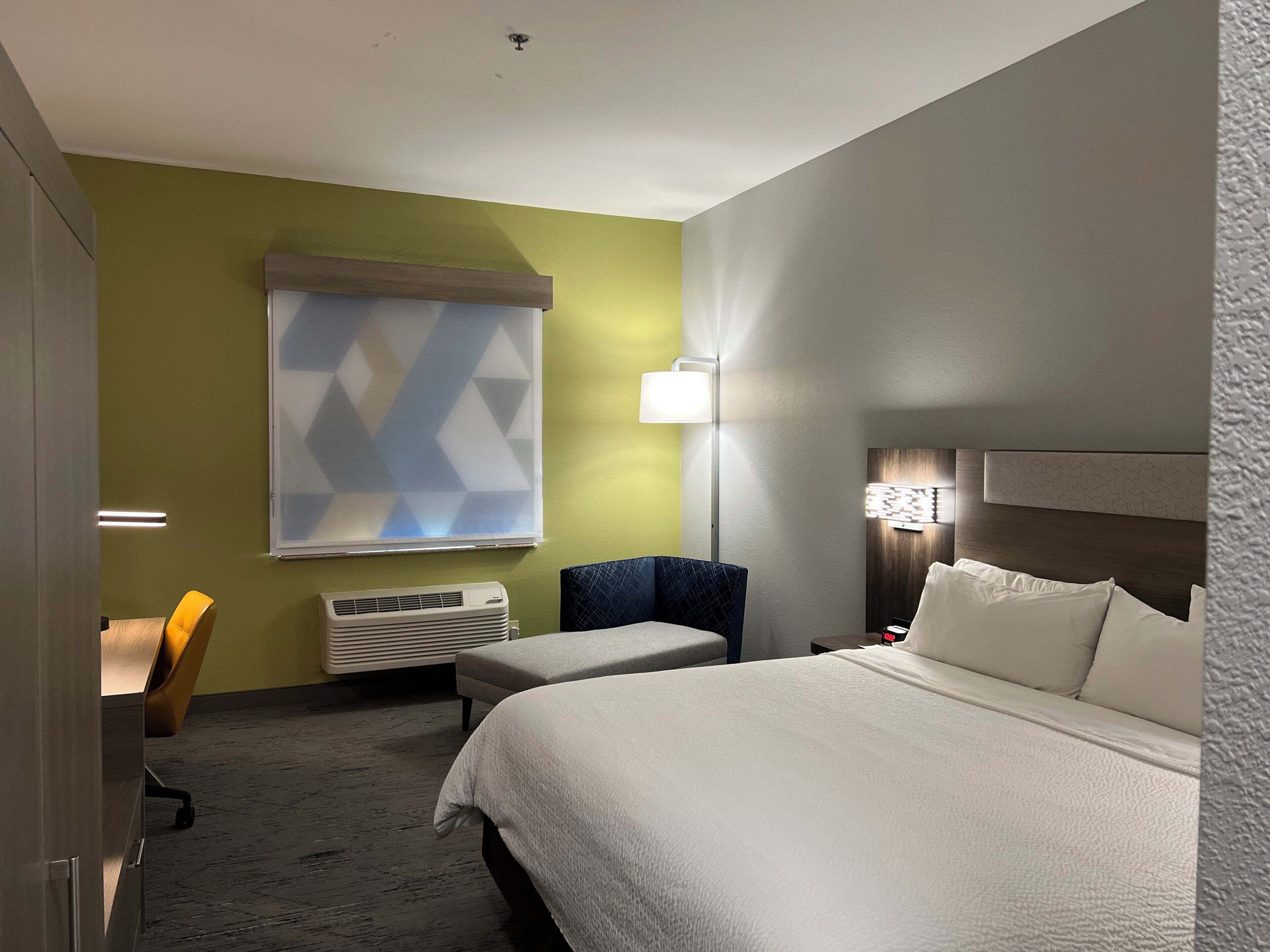 A hotel room with a large bed, a desk, a chair, and a window with a geometric pattern.
