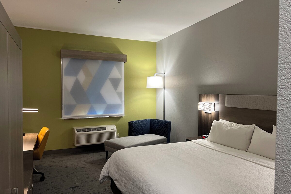 A hotel room with a large bed, a desk, a chair, and a window with a geometric pattern.
