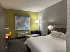 Quarto de hotel com uma cama grande, uma escrivaninha, uma cadeira e uma janela com padrão geométrico.